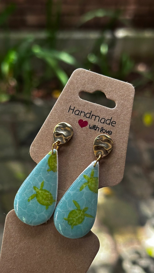 Teardrop-shaped earrings with turtle design on a brown card, held by a hand.